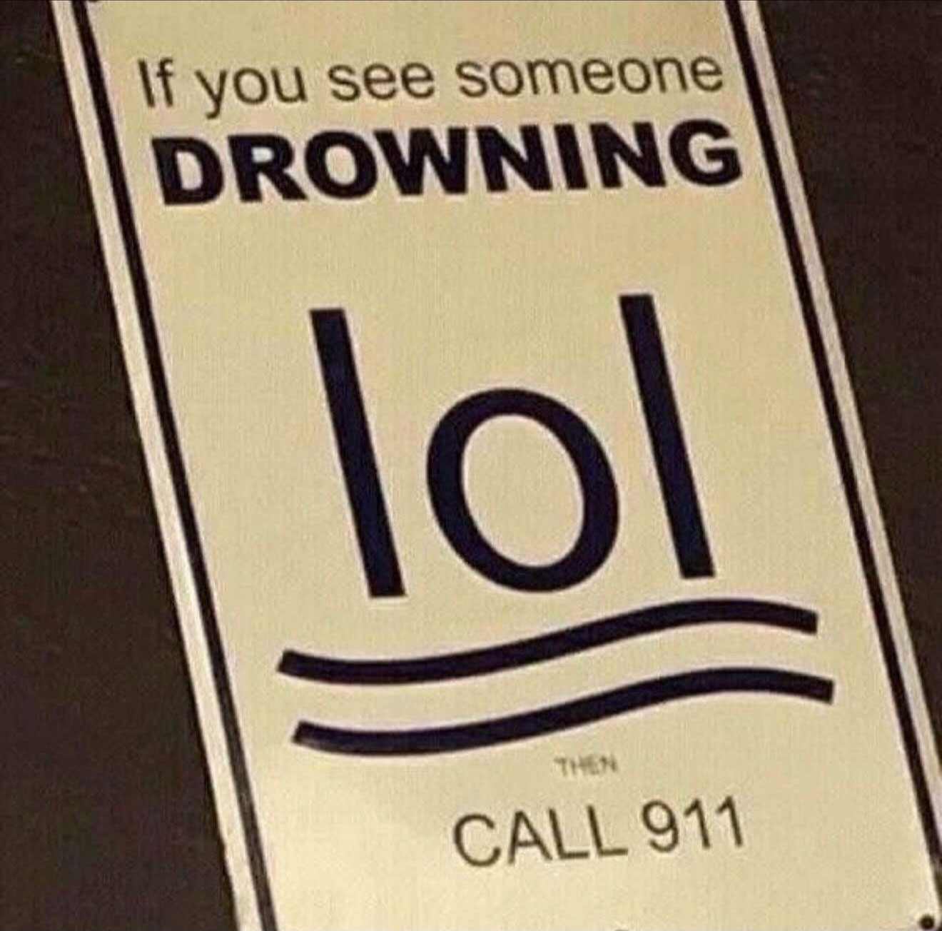A photo of a sign near near water that reads 'If you see someone drowning, then call 911' and to illustrate a person drowning it uses the letters 'lol', as if laughing at the drowning person.
